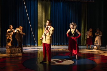 Vilkkaassa näyttämökohtauksessa nähdään näyttelijöitä värikkäissä 1940-luvun puvuissa; mies, jolla on mikrofoni, ja nainen punaisissa hanskoissa seisovat valokeilassa, kun taas muut ovat vuorovaikutuksessa suuren köyden ja taustalla olevien sinisten verhojen lähellä.