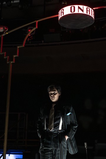 Nastanauhapukuinen henkilö seisoo hehkuvan ON AIR -kyltin alla pimeässä huoneessa, joka on valaistu dramaattisesti alhaalta päin.