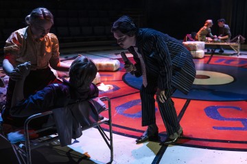 Lavalla on neljä esiintyjää dramaattisen valaistuksen alla. Yksi raidalliseen pukuun pukeutunut henkilö kumartuu eteenpäin ja puhuu mikrofoniin toista, pinnasängyllä istuvaa henkilöä kohti. Kaksi muuta on vuorovaikutuksessa taustalla istuen pinnasängyillä, joissa on huopia.