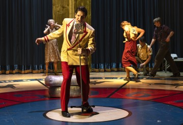 Keltaiseen takkiin ja punaisiin housuihin pukeutunut mies laulaa lavalla vintage-mikrofoniin, kun taas neljä vintage-asuihin pukeutunutta henkilöä näyttelee dramaattisesti taustalla lavavalojen alla.