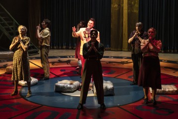 Seitsemän vintage-asuihin pukeutunutta näyttelijää seisoo dramaattisissa asennoissa kirkkaasti valaistulla näyttämöllä, joka on järjestetty suuren, värikkään numeropyörän päälle. Tausta on tumma, ja valonheittimet korostavat esiintyjien ilmeitä.