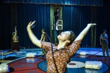 Teatteriesitys lavalla, jossa on viisi näyttelijää; yksi seisoo tikkailla, muut poseeraavat dramaattisesti. Etualalla oleva näyttelijä on hämärtynyt kädet kohotettuna. Lavalla on pyöreitä kuvioita ja käärittyjä esineitä. Taustalla roikkuvat siniset verhot.