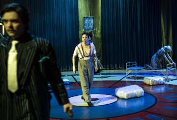 Keskeisellä paikalla seisoo henkselihousuihin pukeutunut henkilö dramaattisessa valaistuksessa, kun taas etualalla kävelee toinen pukuun pukeutunut henkilö. Taustalla joku istuu sairaalasängyllä, jonka lattialla on paketteja, mikä luo jännittyneen tunnelman.