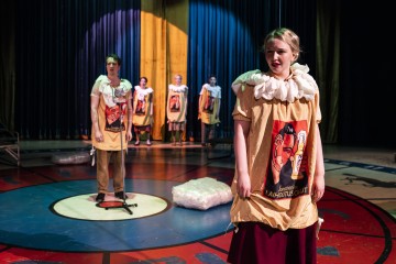 Värikkäisiin pukuihin ja pellekauluksiin pukeutuneet näyttelijät seisovat pyöreällä näyttämöllä, jossa on rohkeita punaisia ja sinisiä kuvioita. Taustalla on siniset verhot ja dramaattinen valaistus, jotka luovat teatteritunnelman.