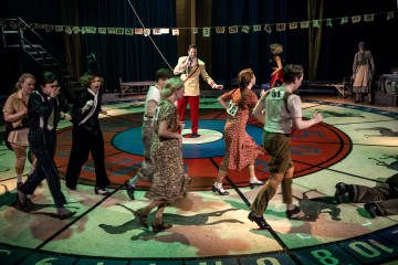 Vintage-vaatteisiin pukeutuneet esiintyjät kävelevät ympyrää värikkäällä numeroidulla näyttämöllä, kun pukuun ja punaisiin housuihin pukeutunut mies seisoo keskellä mikrofonin kanssa; yläpuolella roikkuu lippalakki, ja muut katselevat sivusta.