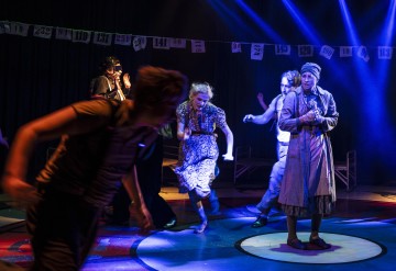 Vintage-vaatteisiin pukeutuneet näyttelijät esiintyvät näyttämöllä energisesti dramaattisten sinisten valojen alla, taustalla roikkuu numeroituja kortteja ja sumea liike viittaa dynaamiseen liikkeeseen.