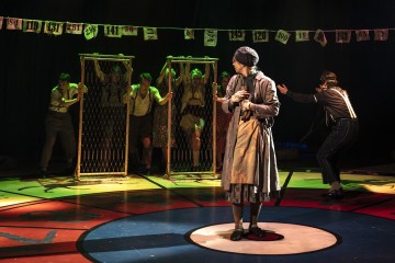 Harmaaseen takkiin ja hattuun pukeutunut henkilö seisoo näyttämön keskellä, kun taas neljä muuta on vuorovaikutuksessa verkkokangasnäyttöjen takana; heidän yläpuolellaan on värikkäitä numeroituja kortteja, ja näyttämön lattialla on rohkea pyöreä kuvio. Valaistus on dramaattinen.