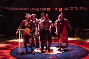 Viisi näyttelijää seisoo yhdessä kirkkaasti valaistulla näyttämöllä, pukeutuneina vintage-tyylisiin vaatteisiin ja katsoen ylöspäin. Heidän takanaan roikkuu numeroituja lippuja ja keltainen aita, ja dramaattinen valaistus luo teatteritunnelmaa.