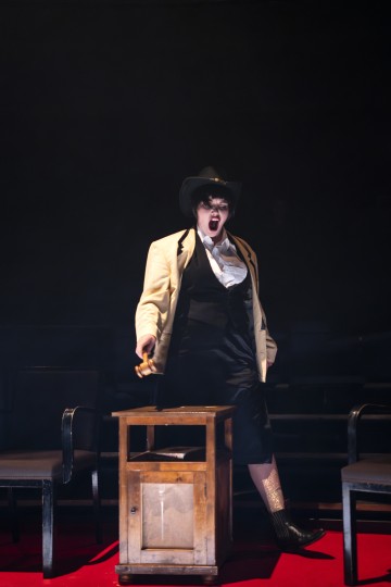 Kermanväriseen takkiin, mustaan liiviin, hameeseen, cowboy-hattuun ja saappaisiin pukeutunut esiintyjä seisoo lavalla puulaatikon päällä, suu dramaattisesti auki, ja valokeila korostaa häntä tummaa taustaa vasten.