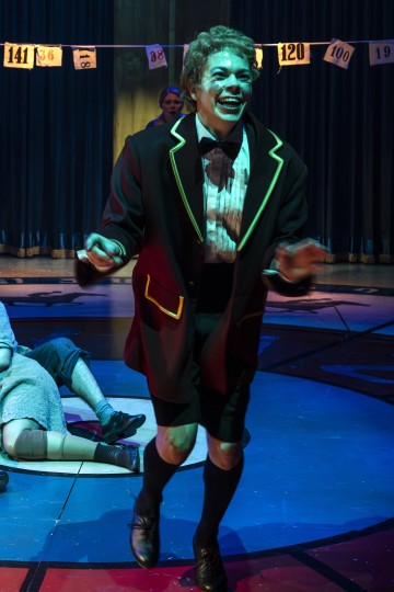 Henkilö, jolla on tumma bleiseri, jossa on keltainen reunus, shortsit ja polvisukat, tanssii iloisesti värikkäällä lavalla. Taustalla lattialla makaa muita, ja yläpuolella roikkuu numeroituja banderolleja.