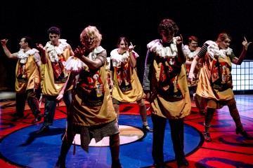 Ryhmä esiintyjiä värikkäissä puvuissa, joissa on liioitellut kaulukset ja esiliinat, poseeraa dramaattisesti kirkkaasti valaistulla näyttämöllä, jossa on rohkeita punaisia ja sinisiä lattiakuvioita.