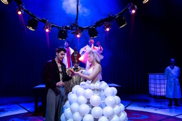 Värikkäästi valaistu teatterikohtaus, jossa on mies ja nainen puvuissa, naisella valkoisista ilmapalloista tehty hame. Heidän takanaan seisoo kolme valkoisiin pukeutunutta esiintyjää, ja sivummalla seisoo valkoiseen laboratoriotakkiin pukeutunut henkilö.