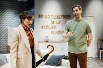 Sateenvarjo kädessä oleva, yllättyneen näköinen nainen seisoo vihreään paitaan pukeutuneen miehen vieressä, joka elehtii kesken keskustelun. Heidän takanaan seinällä on kyltti, jossa lukee: BEING TOGETHER IS AN ENDLESS SUNDAY.