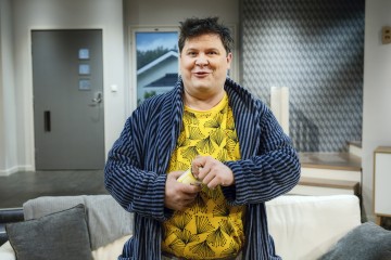 Mies, jolla on sininen raidallinen kaapu kirkkaankeltaisen kuvioidun paidan päällä, seisoo sisätiloissa hymyillen ja pitäen jotakin käsissään, ja taustalla on harmaa sohva ja moderni sisustus.