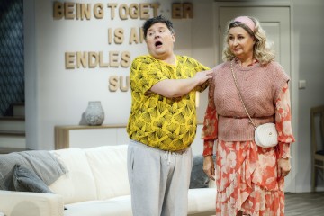 Mies, jolla on keltainen kuvioitu paita ja harmaat housut, tekee eleitä seisoessaan vaaleanpunaiseen kukka-asuun ja päähineeseen pukeutuneen naisen vieressä. He ovat olohuoneessa, jossa on sohva ja heidän takanaan seinäkyltti.