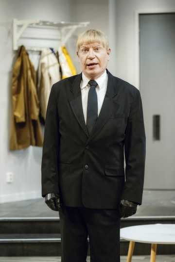 Vaaleahiuksinen mies, jolla on musta puku, solmio ja käsineet, seisoo sisätiloissa vaatetelineen ja oven edessä ja puhuu tai esiintyy ilmeikkäästi.