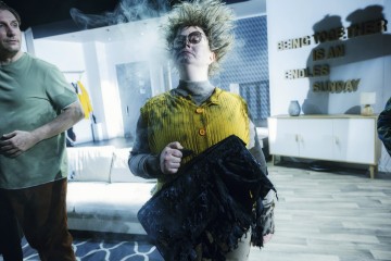 Sotkuinen tukkainen ja silmälasipäinen henkilö, jolla on keltainen liivi, seisoo sisätiloissa pitäen kädessään palanutta salkkua. Salkusta nousee savua. Seinällä olevassa kyltissä lukee: YHDESSÄ OLEMINEN ON LOPUTON SUNNUNTAI.