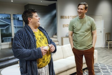 Kaksi miestä seisoo ja puhuu olohuoneessa. Toisella on raidallinen kaapu ja keltainen paita, ja hänellä on kuppi kädessään. Toinen, vihreä t-paita ja ruskeat housut, hymyilee. Taustalla on sohva ja seinätaidetta.