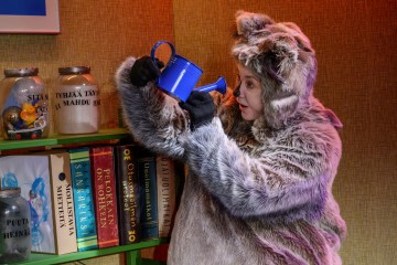 Karvaiseen eläinasuun pukeutunut henkilö pitää kädessään sinistä kastelukannua ja katselee sitä uteliaana. Heidän takanaan on kirjoja hyllyllä ja suuria purkkeja täynnä pieniä esineitä. Kohtaus on kirkkaasti valaistu ja leikkisä.