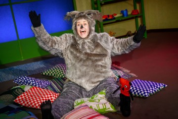 Henkilö, jolla on pörröinen harmaa eläinasu, istuu lattialla kädet ylhäällä ja hymyilee värikkäiden kuvioitujen tyynyjen ympäröimänä kirkkaasti valaistussa huoneessa, jossa on vihreät seinät ja sininen valaistus.