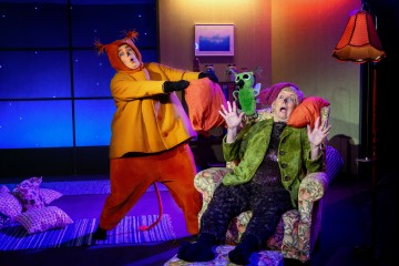 Oranssiin pukuun pukeutunut henkilö, jolla on antennit, osoittaa dramaattisesti istuvaa, yllättynyttä henkilöä, joka pitää vihreää nukkea nojatuolissa, kirkkaasti valaistulla näyttämöllä, jossa on värikkäitä tyynyjä ja lamppu.