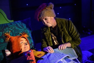 Kaksi näyttelijää näyttämöllä: toinen makaa sängyssä oranssit eläimen korvat ja nenä maalattuna, kun taas toinen, jolla on neuletakki ja vihreä takki, nojaa heidän ylleen. Kohtaus on valaistu sinisellä valaistuksella, joka luo yöllisen tunnelman.