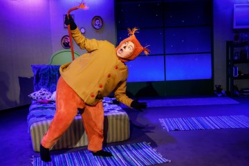 Oranssiin pukuun pukeutunut esiintyjä, jolla on häntä ja tupsullinen päähine, esiintyy näyttämöllä dramaattisesti seisoen sängyn vieressä huoneessa, jonka seinät ja matot ovat siniset, mikä herättää mielikuvituksen teatterikohtauksesta.