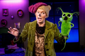 Karhupukuun pukeutunut esiintyjä, jolla on ruskea hattu ja maalattu nenä, pitää lavalla vihreää nukkea, jolla on suuret silmät ja räpyttelevät korvat, värikkäällä, lämpimästi valaistulla taustalla.