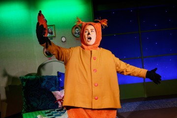 Oranssiin pukuun pukeutunut henkilö, jolla on teräväkorvaiset korvat ja tupsut, nostaa pientä eläintä muistuttavaa käsinukkea, joka seisoo värikkäässä teatterihuoneessa, jossa on vihreä seinä ja sininen tähtitausta.