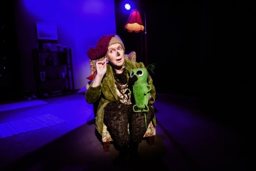 Värikkäisiin, kodikkaisiin vaatteisiin ja hassuun hattuun pukeutunut henkilö istuu nojatuolissa hämärästi valaistulla näyttämöllä, pitelee vihreää, pehmoista nukkea ja elehtii ilmeikkäästi valokeilan alla.