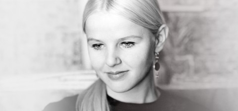 Mustavalkoinen valokuva nuoresta naisesta, jolla on taaksepäin vedetyt vaaleat hiukset ja korvakorut, ja joka katsoo alaspäin lempeästi hymyillen. Tausta on pehmeästi sumea.