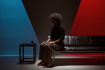 Kiharatukkainen henkilö istuu mustalla sohvalla hämärässä huoneessa, jota valaisee sininen valo vasemmalla ja punainen valo oikealla. Sohvan vieressä on pieni pöytä, jossa on lasi.