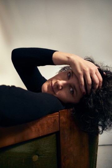 Tummahiuksinen nainen hymyilee vienosti ja pitelee otsaansa. Hän makaa jonkinlaisella huonekalulla.