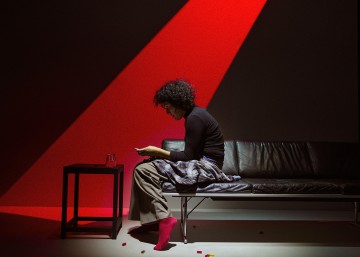 Kiharatukkainen henkilö istuu mustalla sohvalla dramaattisen punaisen valonheittimen alla ja katsoo puhelinta. Hänellä on punaiset sukat ja ruudulliset housut, ja värillisiä palikoita on ripoteltu lattialle ja pienelle pöydälle hänen vieressään.