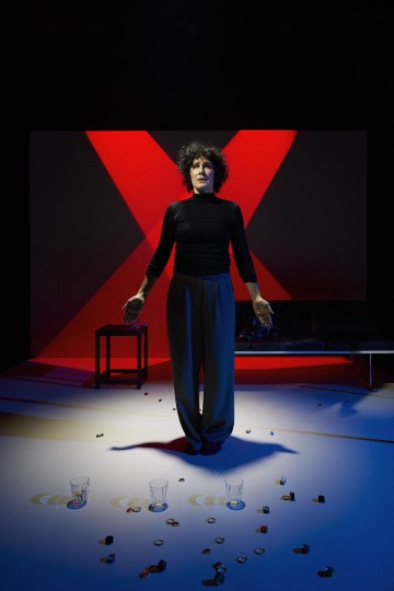 Nainen seisoo näyttämöllä edessään kolme juomalsia ja pullonkorkkeja. Taustalla on pieni pöytä ja nahkasohva