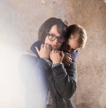 Silmälasipäinen nainen halaa lasta tiiviisti, molemmat näyttävät vakavilta ja synkiltä, ja taustalla on neutraali tausta, jossa on pehmeä valo vasemmalla.