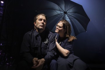 Tummapukuinen mies istuu silmät kiinni, kun hymyilevä nainen hänen vieressään pitää avattua sateenvarjoa heidän yläpuolellaan ja hänen kätensä lepää miehen olkapäällä, ja molempia valaisee dramaattinen näyttämövalaistus.