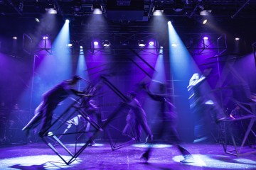 Esiintyjät liikkuvat energisesti lavalla suurten metallikehysten kanssa dramaattisessa sinisessä ja violetissa valaistuksessa, joka luo dynaamista liikesumua; valonheittimet loistavat ylhäältä ja korostavat teatterin tunnelmaa.