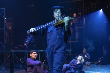 Siniseen haalariin ja huiviin pukeutunut henkilö soittaa lavalla viulua, ja muut samankaltaisiin asuihin pukeutuneet istuvat tai lepäävät heidän ympärillään dramaattisessa valaistuksessa.