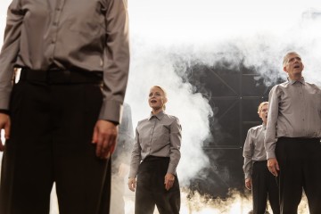 Joukko harmaisiin paitoihin ja mustiin housuihin pukeutuneita ihmisiä seisoo savuisen taustan edessä, ja joillakin heistä on suu auki, ikään kuin he laulaisivat tai puhuisivat. Kohtaus vaikuttaa dramaattiselta ja lavastetulta.