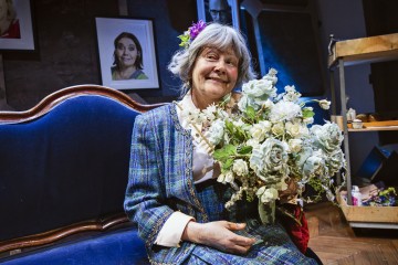 Iäkäs harmaahiuksinen nainen, jolla on siniruutuinen asu ja kukka hiuksissaan, istuu hymyillen sinisellä sohvalla ja pitää kädessään suurta kimppua vaaleansinisiä ja valkoisia kukkia. Taustalla näkyy kehystettyjä muotokuvia ja kärry.