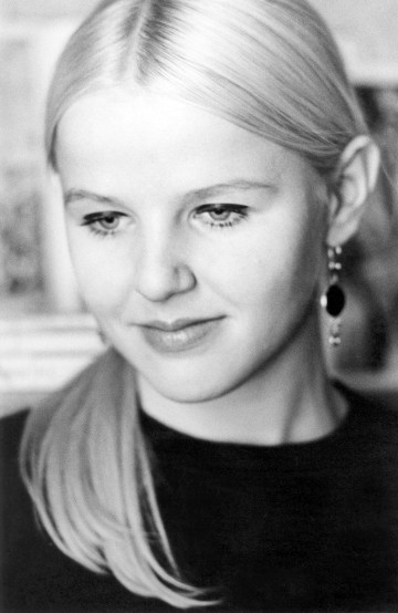 Mustavalkoinen muotokuva nuoresta naisesta, jolla on suorat, vaaleat, keskeltä erotetut hiukset, tumma toppi ja roikkuvat korvakorut, ja joka katsoo alaspäin lempeällä, mietteliäällä ilmeellä.