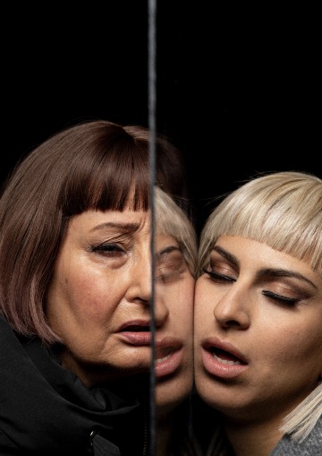 Kaksi lyhythiuksista naista, toinen vanhempi ja toinen nuorempi, seisovat tiiviisti yhdessä mustaa taustaa vasten. Pystysuora peili jakaa heidän kasvonsa ja sulauttaa heidän heijastuksensa ja piirteensä yhdeksi tunteikkaaksi kuvaksi.