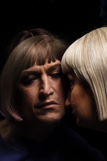 Henkilö, jolla on lyhyet, vaaleanruskeat hiukset, katsoo vakavasti peilikuvaansa peilistä, mikä luo illuusion kahdesta vastakkain olevasta ihmisestä pimeässä, dramaattisessa ympäristössä.