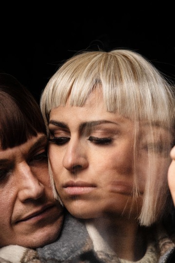 Lähikuva kahdesta lyhythiuksisesta ihmisestä: toisella on vaaleat vaaleat hiukset ja otsatukka, toisella ruskeat hiukset. Heidän kasvonsa ovat painautuneet yhteen, osittain sumeat, mikä viittaa läheisyyteen ja emotionaaliseen syvyyteen.