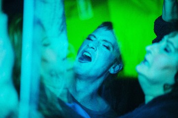 Henkilö laulaa intohimoisesti suu auki eloisan sinisen ja vihreän valaistuksen keskellä, muiden ympäröimänä, mikä viittaa vilkkaaseen, energiseen ilmapiiriin, kuten konserttiin tai juhliin.