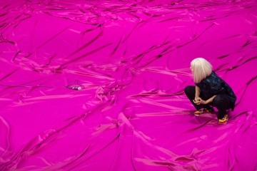 Valkohiuksinen henkilö kyykistyy suurelle, kirkkaan vaaleanpunaisella kankaalla päällystetylle pinnalle ja katselee pientä laitetta, jonka kaapelit makaavat lähellä kankaan päällä.