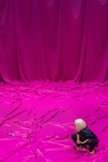 Platinanvaaleat hiukset omaava henkilö kyykistelee lattialla, jota peittää rypistynyt, kirkkaan magentanpunainen kangas, joka sopii yhteen verhojen taustan kanssa. Mikrofoni ja johto makaavat kankaan päällä lähellä.