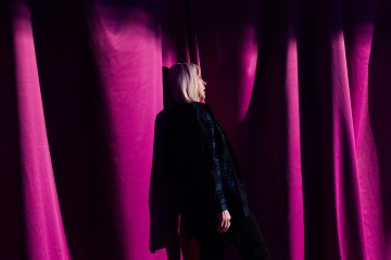 Vaalea vaaleatukkainen henkilö seisoo dramaattisten, syvän vaaleanpunaisten verhojen edessä, joita osittain valaisee läpi virtaava auringonvalo, joka luo kankaaseen rohkeita varjoja ja kohokohtia.