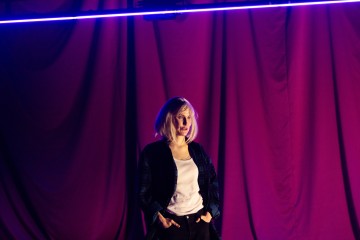 Henkilö, jolla on hartioiden pituiset vaaleat hiukset, seisoo violettien verhojen edessä, joita valaisee sininen neonvalo. Hänellä on yllään musta takki ja valkoinen paita, kädet taskuissa, katse hieman sivulle.